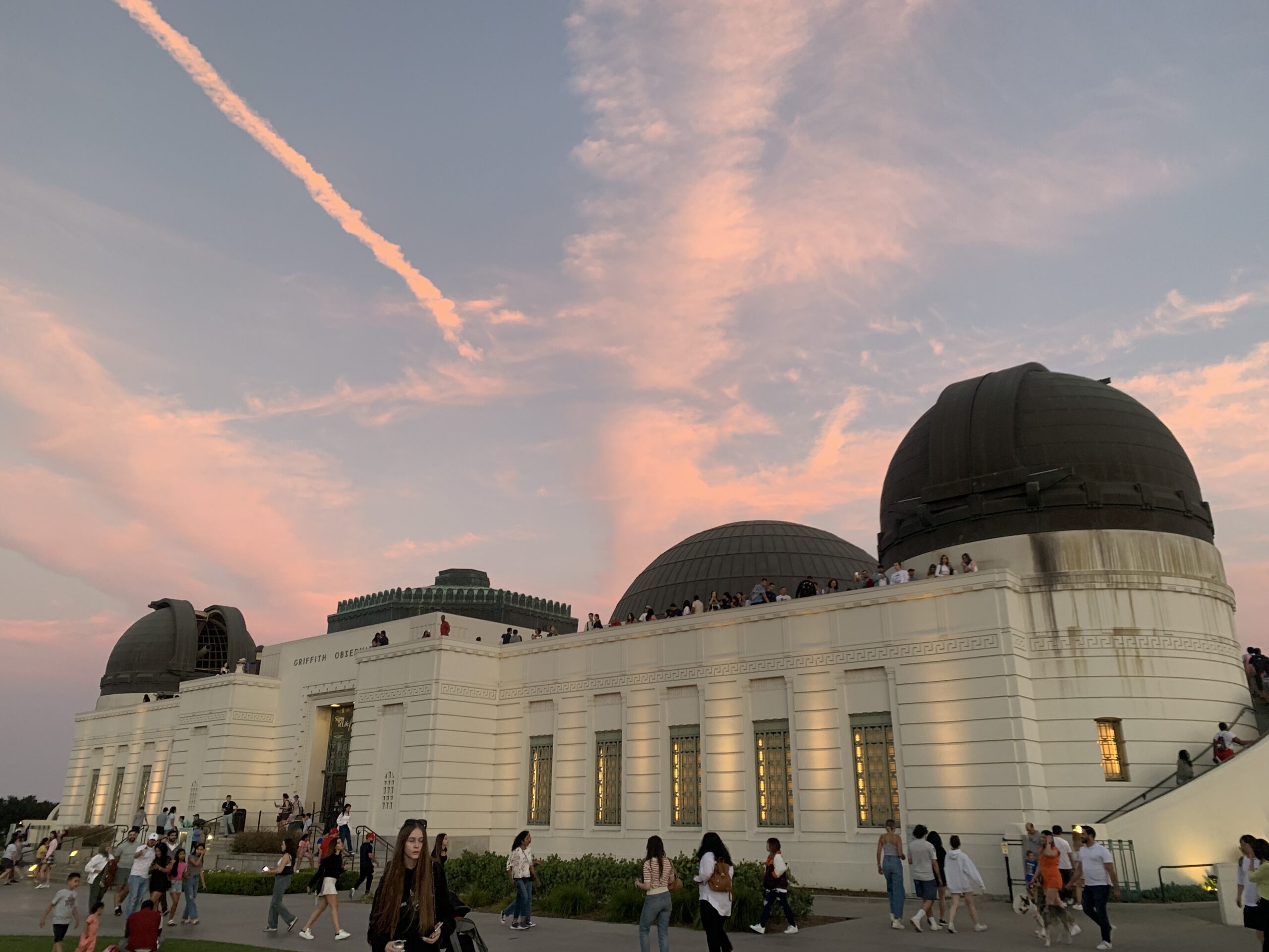 The front facade of Griffith Observatory on a busy summer day with many visitors on the roof and front lawn. Just after sunset, pink clouds are above the building in the background.