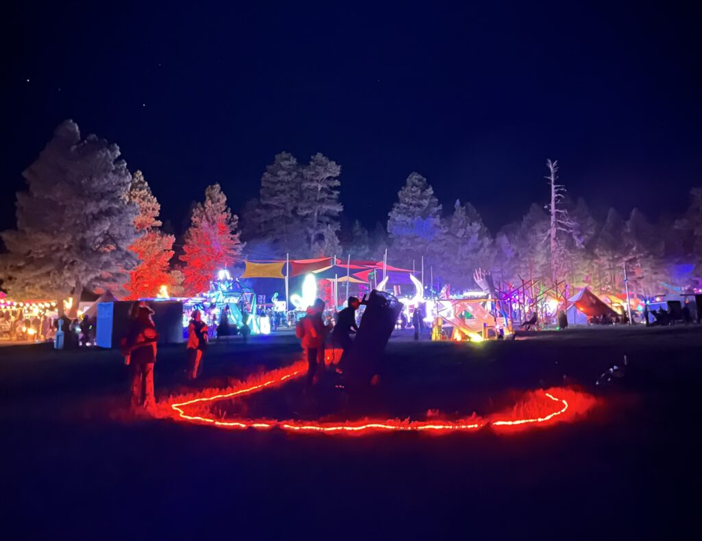 Katie adjusting the 20" telescope at Desert Hearts Festival 2024 with glowing art cars in the background (credit Brian Smith).